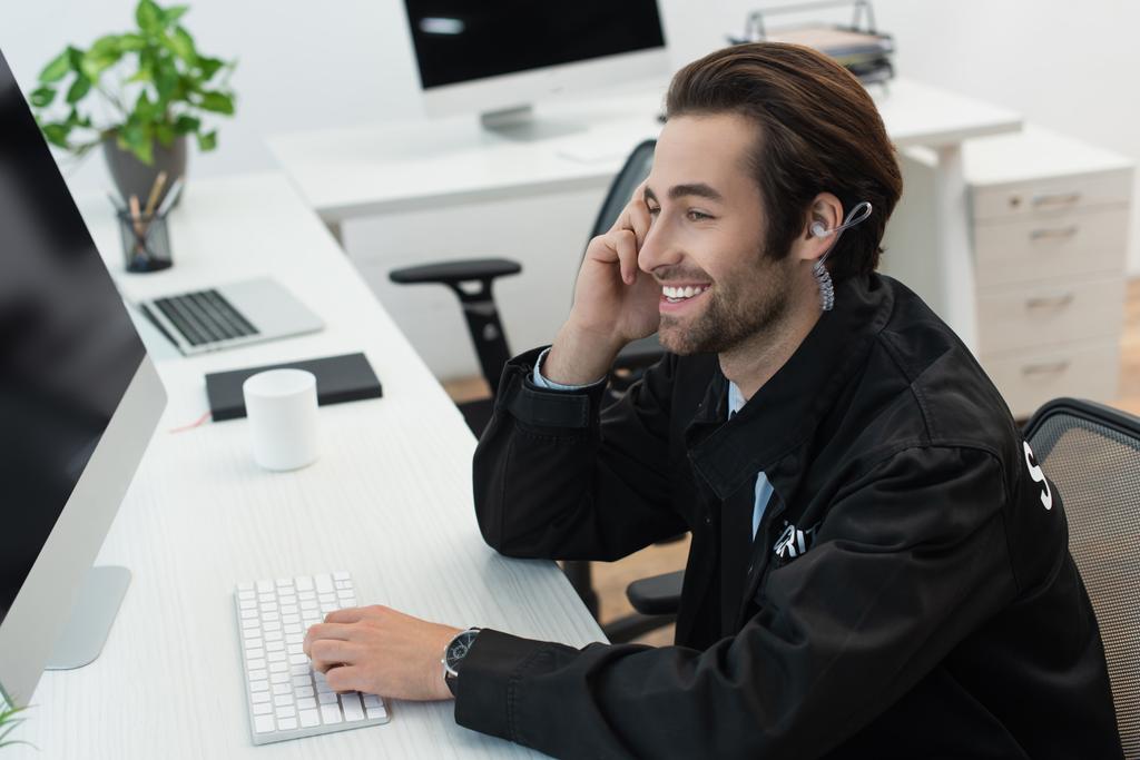 young security man in earphone smiling at workplace near computer monitor - Photo, Image