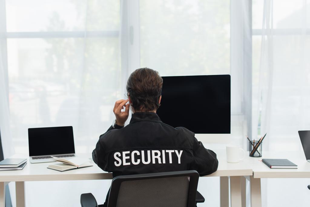 vista posteriore dell'uomo di sicurezza in uniforme nera e auricolare seduto vicino ai computer nella sala di supervisione - Foto, immagini