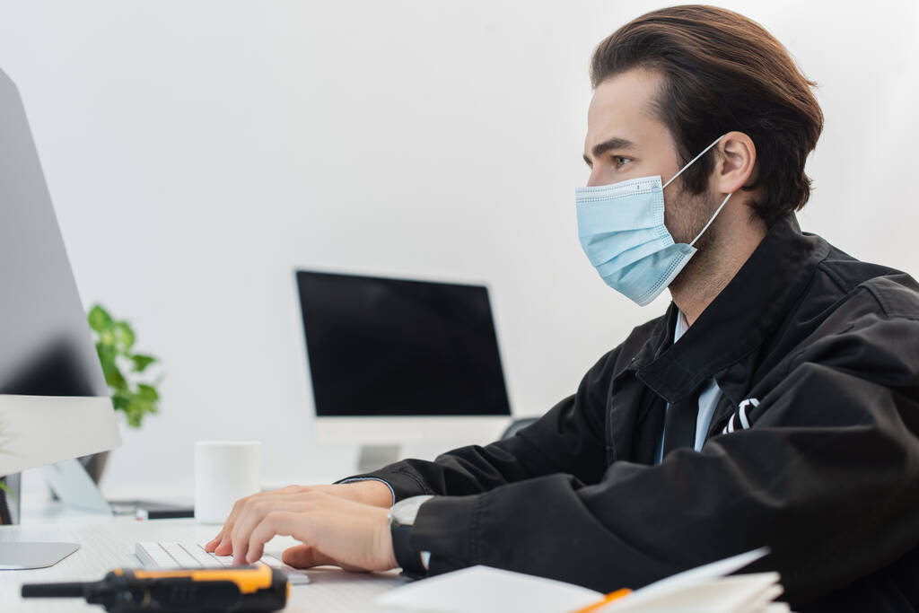 vue latérale du gardien dans un masque médical travaillant sur un ordinateur près du talkie-walkie flou - Photo, image