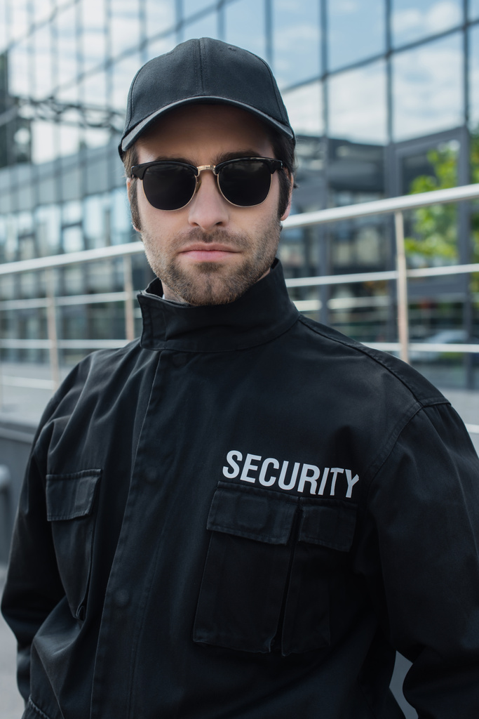 Siyah üniformalı ve güneş gözlüklü bir güvenlik görevlisi dışarıdaki kameraya bakıyor. - Fotoğraf, Görsel