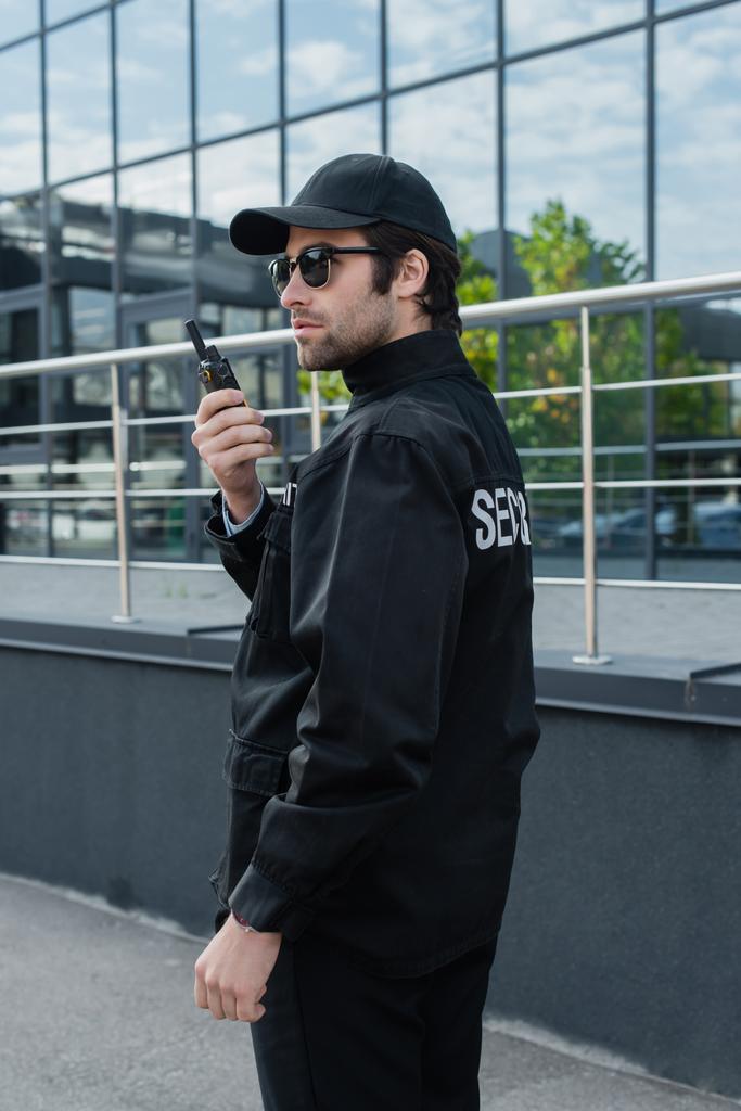joven hombre de seguridad en uniforme, gorra y gafas de sol hablando en walkie-talkie cerca del edificio - Foto, Imagen