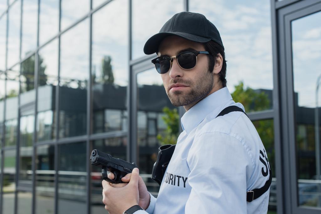 Siyah şapkalı ve güneş gözlüklü bir güvenlik görevlisi dışarıda silah tutuyor. - Fotoğraf, Görsel