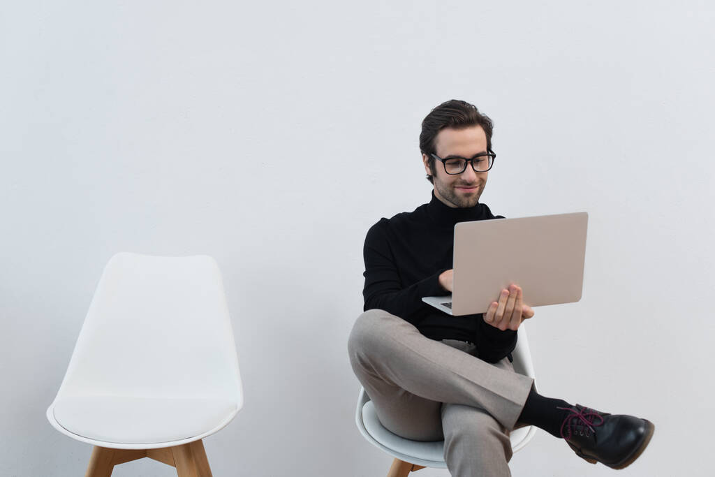 hombre feliz y elegante usando el ordenador portátil mientras está sentado en la silla cerca de la pared gris - Foto, imagen