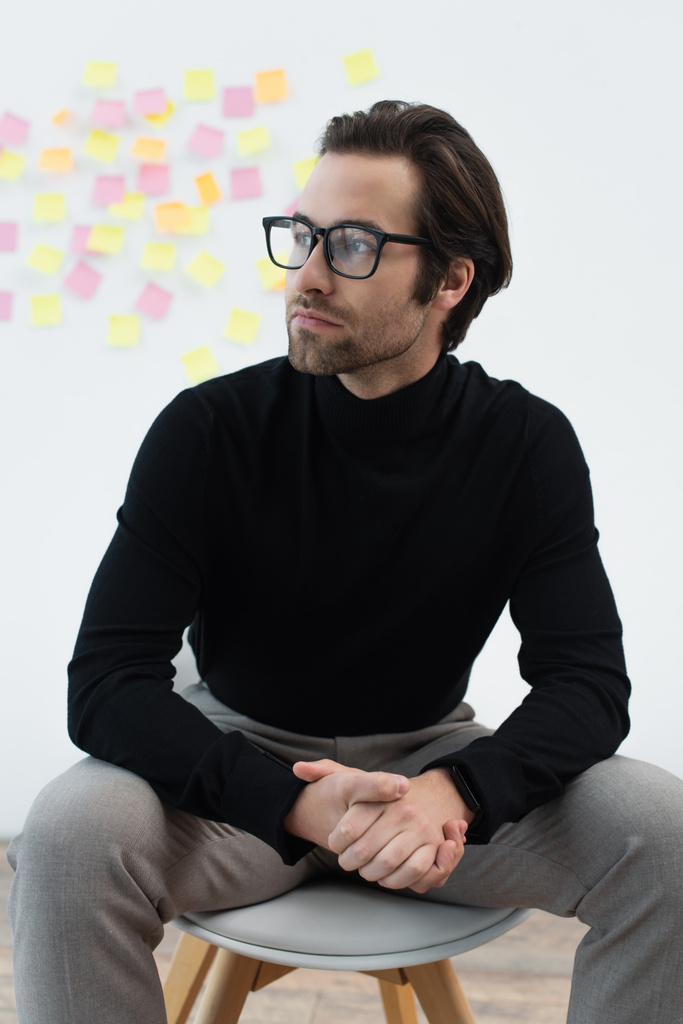 young man in black turtleneck and eyeglasses sitting with clasped hands and looking away - Photo, Image