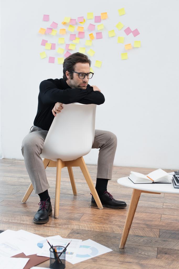 вдумливий чоловік сидить на стільці біля зошитів на кавовому столі і папері на підлозі
 - Фото, зображення