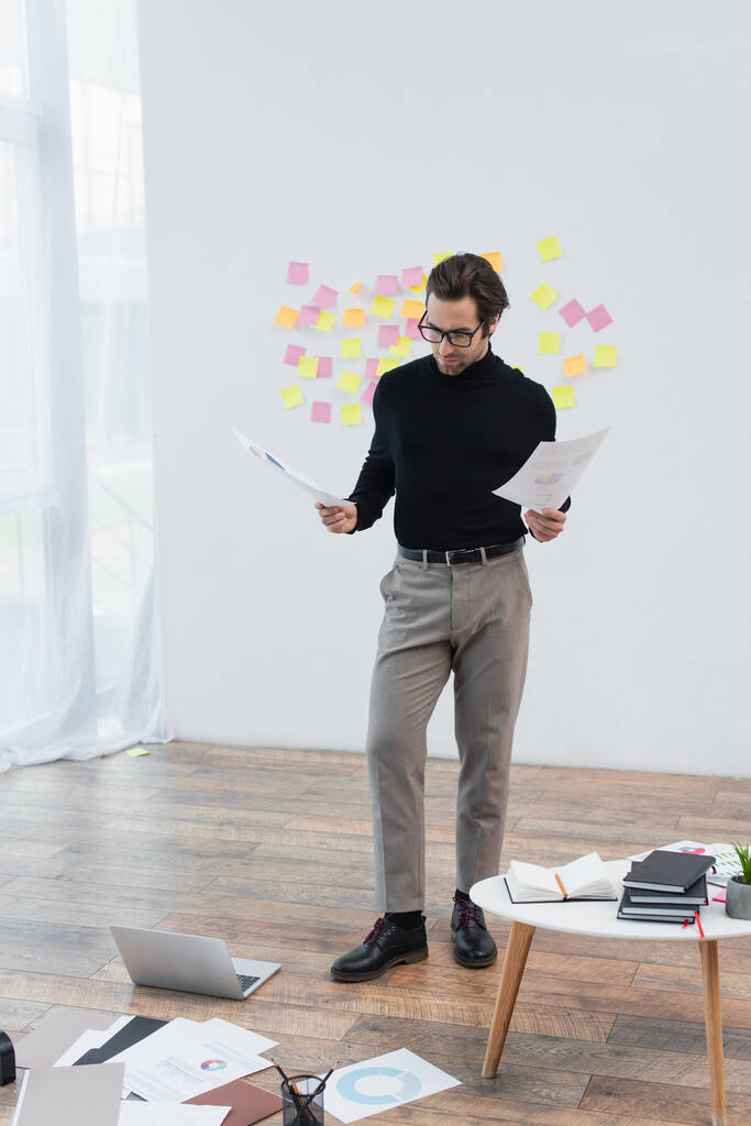 stylový muž s dokumenty stojí v blízkosti notebooku a papíry na podlaze - Fotografie, Obrázek