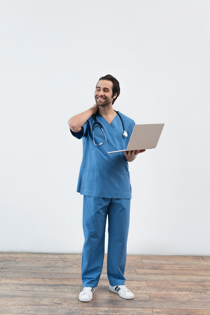 πλήρες μήκος του χαμογελαστού γιατρού με φορητό υπολογιστή κοιτάζοντας μακριά κοντά σε γκρίζο τοίχο - Φωτογραφία, εικόνα