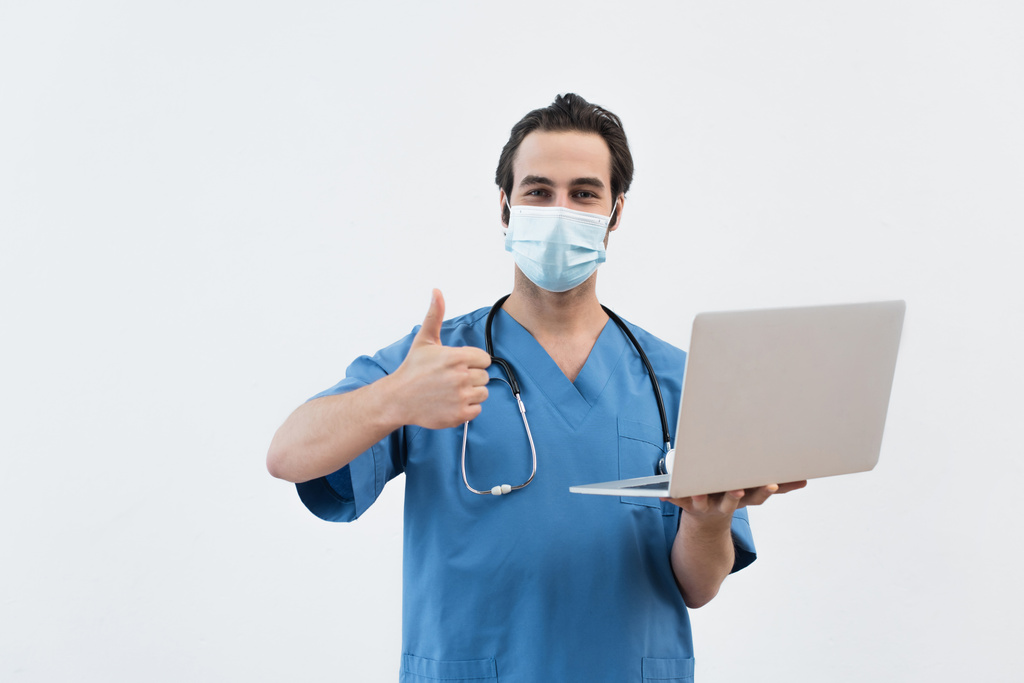 ノートパソコンを持った医療用マスクの若い医者はグレーで隔離されたジェスチャーのように - 写真・画像