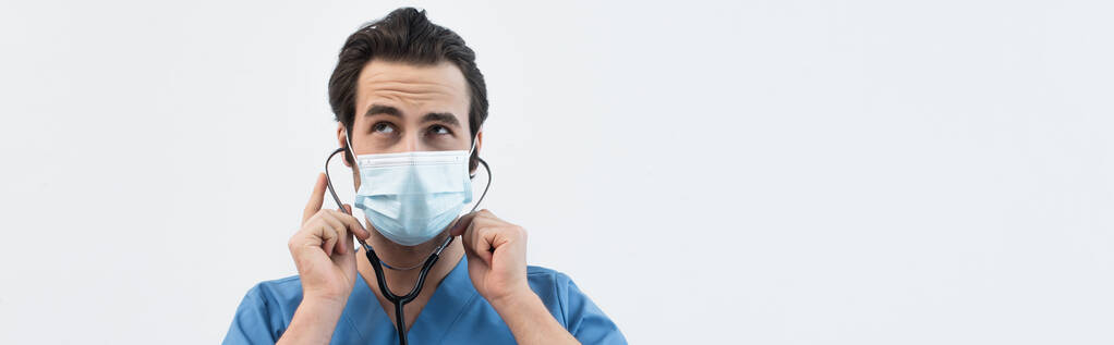 вдумчивый врач в медицинской маске, используя стетоскоп, изолированный на сером, баннер - Фото, изображение