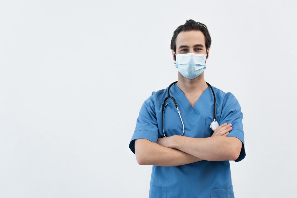 mladý lékař v lékařské masce stojící se zkříženýma rukama a dívající se na kameru izolovanou na šedi - Fotografie, Obrázek