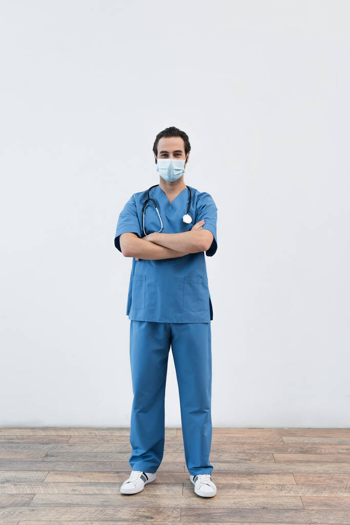 intera lunghezza del medico in maschera medica in piedi con braccia incrociate vicino alla parete grigia - Foto, immagini