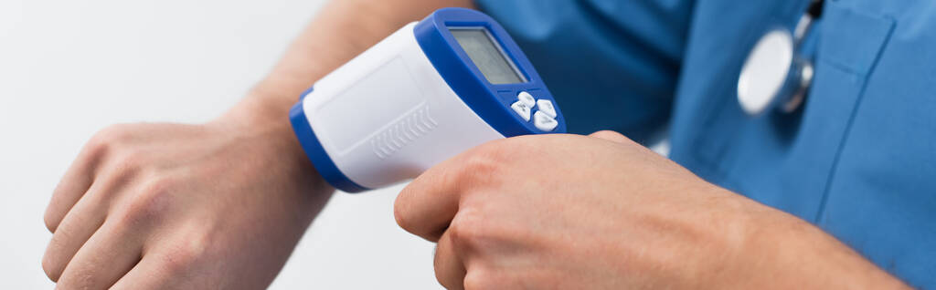 osittainen näkymä hämärtyneestä lääkäristä, joka mittaa omaa lämpötilaansa kosketuksettomalla lämpömittarilla eristettynä harmaalla, bannerilla - Valokuva, kuva