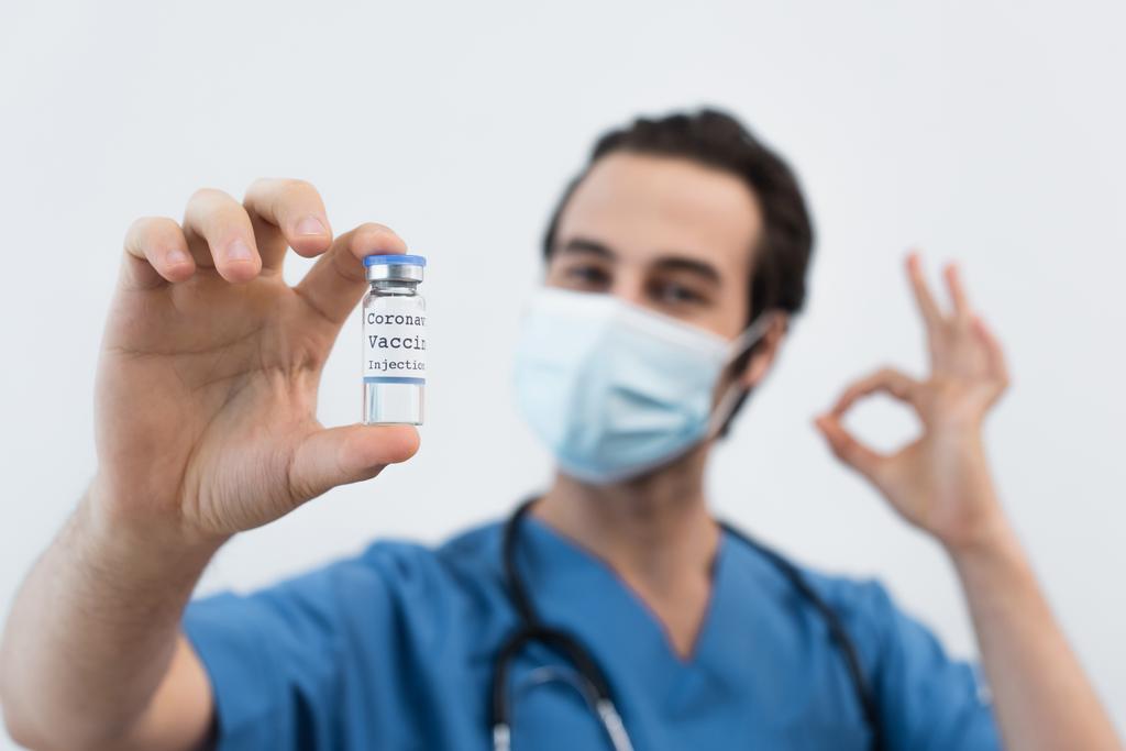 rozmazaný lékař v lékařské masce obsahující koronavirovou vakcínu a ukazující ok gesto izolované na šedi - Fotografie, Obrázek