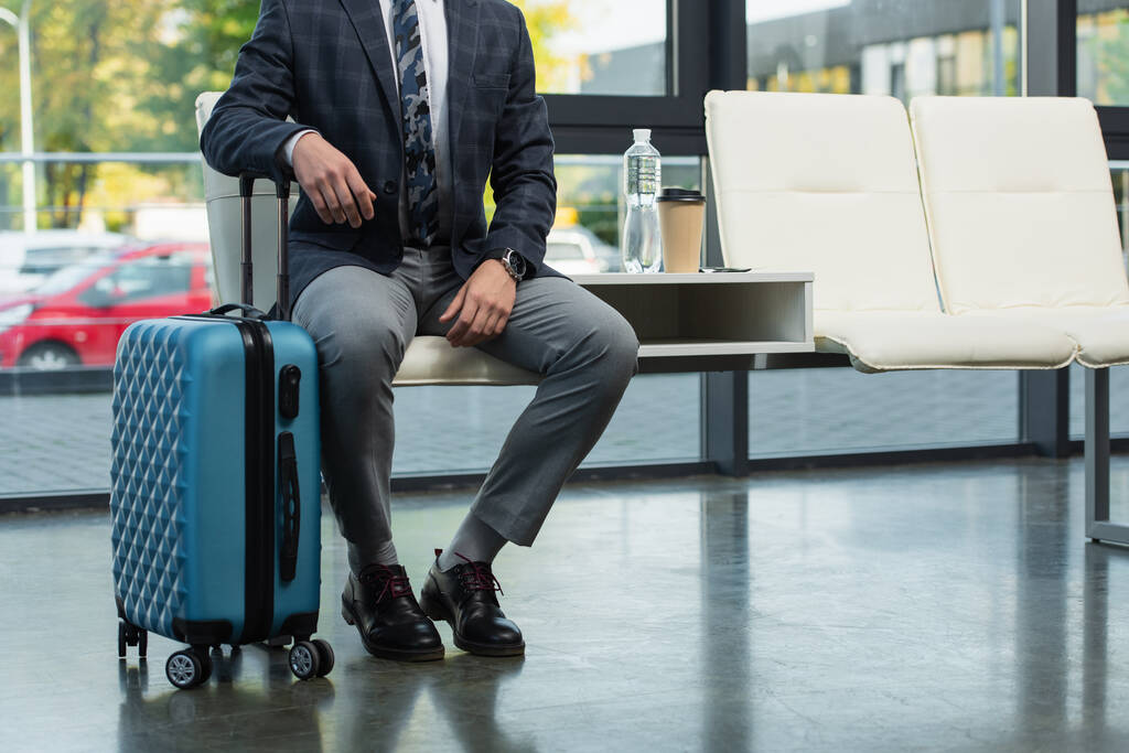 обрезанный вид на путешественника, сидящего в зале вылета рядом с чемоданом, бутылка воды и бумажная чашка - Фото, изображение