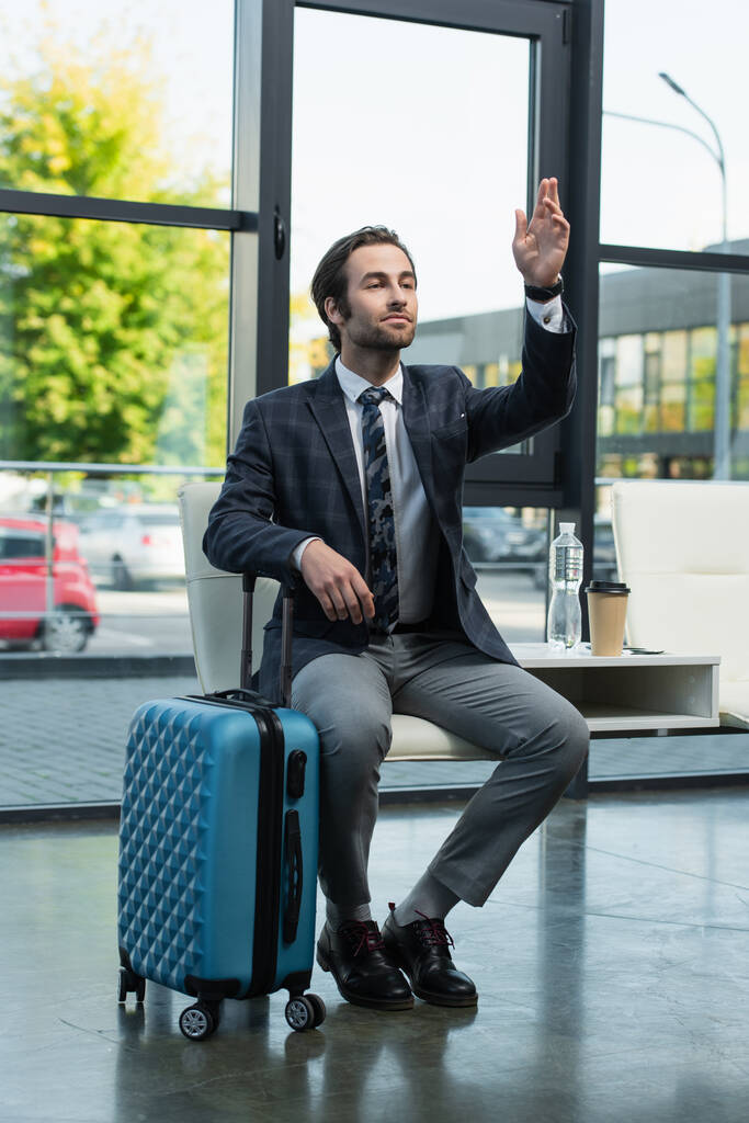 hombre mirando hacia otro lado y saludando la mano mientras espera en la sala de salida cerca de la bolsa de viaje - Foto, Imagen