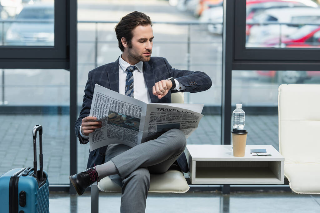 Seyahat hayatı gazetesi olan bir adam kalkış salonunda otururken kol saatine bakıyor. - Fotoğraf, Görsel