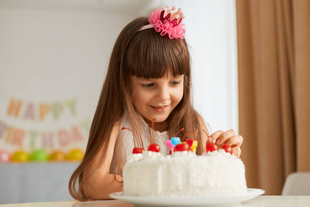 Χαρούμενο ενθουσιασμένο θηλυκό παιδί με σκούρα μαλλιά που διακοσμεί την τούρτα γενεθλίων του, περιμένει γιορτές, ποζάρει στο εορταστικό δωμάτιο μόνη της, το παιδί έχει καλή διάθεση. Χρόνια πολλά.. - Φωτογραφία, εικόνα