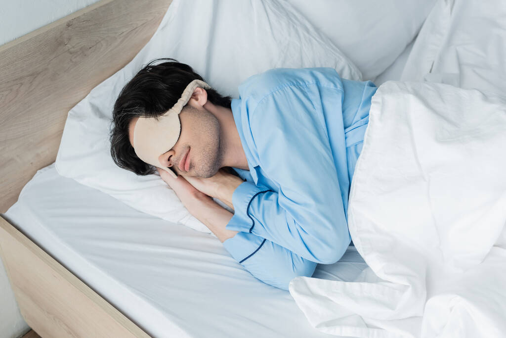 junger Mann in Schlafmaske und Schlafanzug, schlafend auf weißem Bettzeug - Foto, Bild