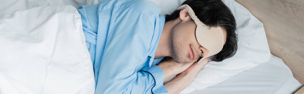νεαρός άνδρας κοιμάται σε λευκό κρεβάτι με μπλε πιτζάμες και μάσκα ύπνου, πανό - Φωτογραφία, εικόνα