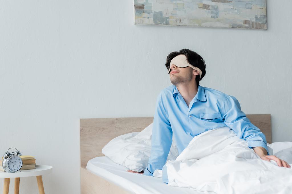 muž ve spánkové masce sedí na posteli v blízkosti retro budík na nočním stolku - Fotografie, Obrázek