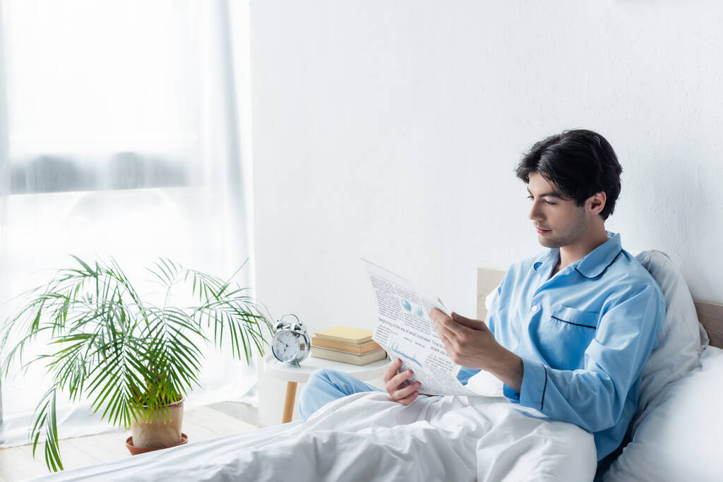 νεαρός άνδρας με μπλε πιτζάμες διαβάζει εφημερίδα κοντά σε παλιό ξυπνητήρι στο κομοδίνο - Φωτογραφία, εικόνα