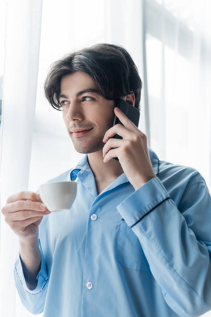sorrindo homem de pijama azul segurando xícara de café enquanto fala no telefone celular - Foto, Imagem