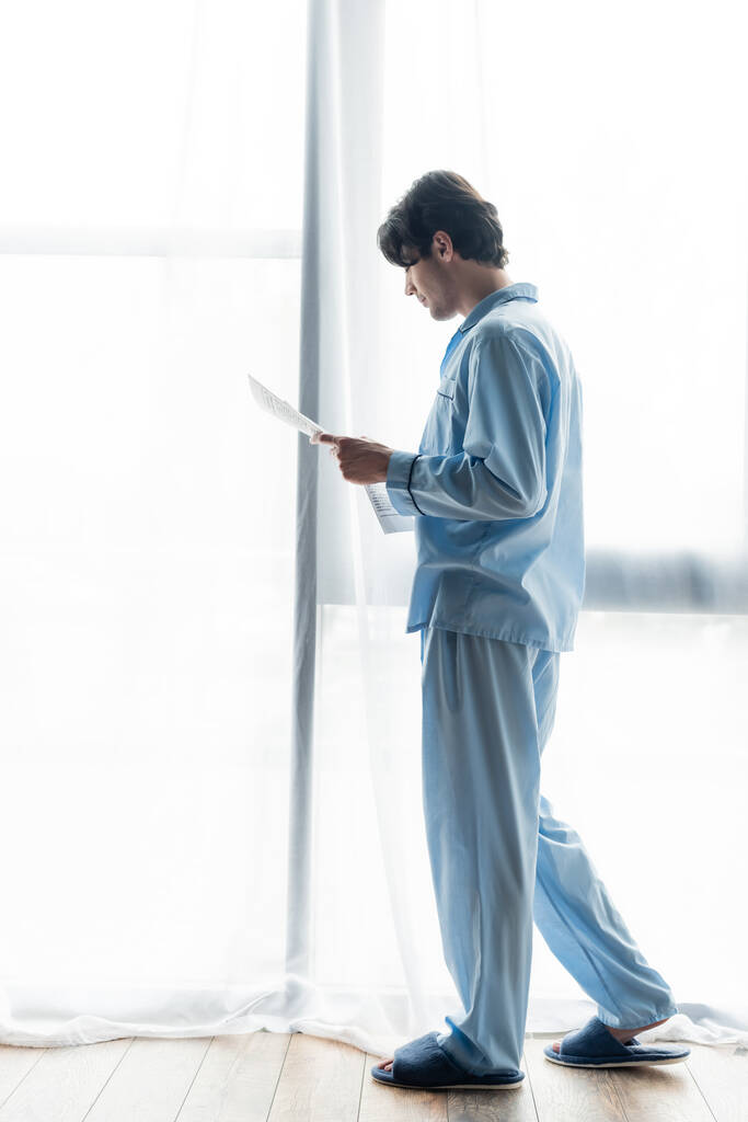 visão de comprimento total do jovem em pijama azul ler jornal perto da janela - Foto, Imagem
