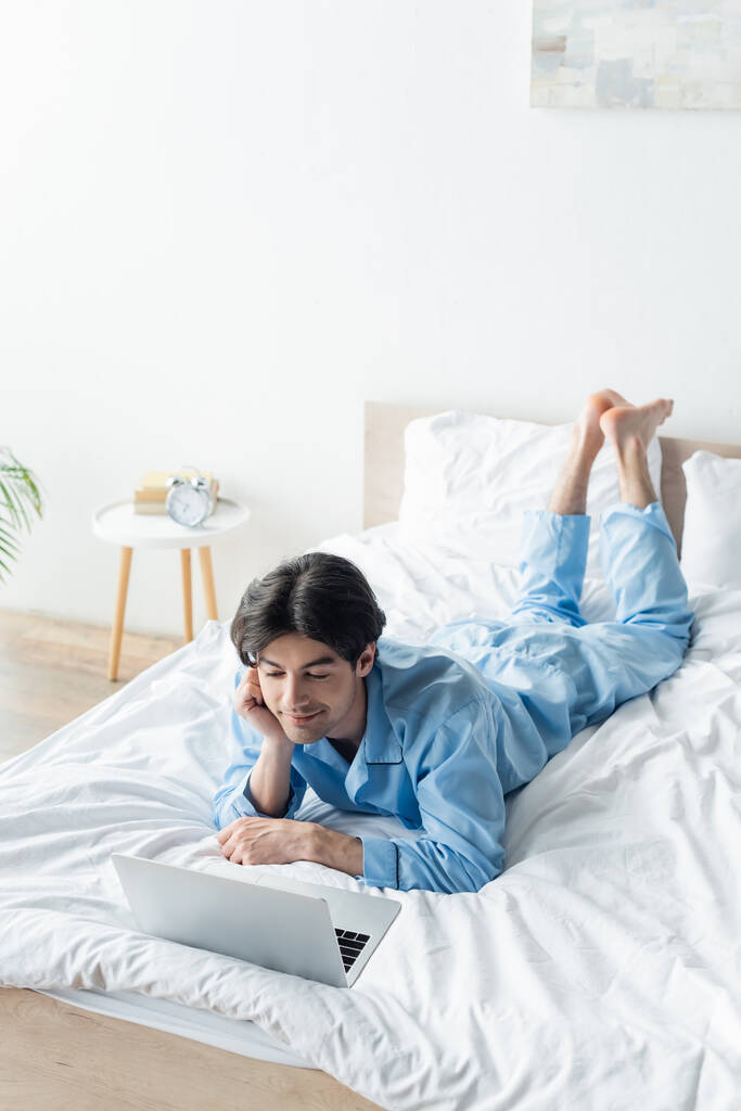 усміхнений чоловік на блакитній піжамі дивиться фільм на ноутбуці на ліжку
 - Фото, зображення