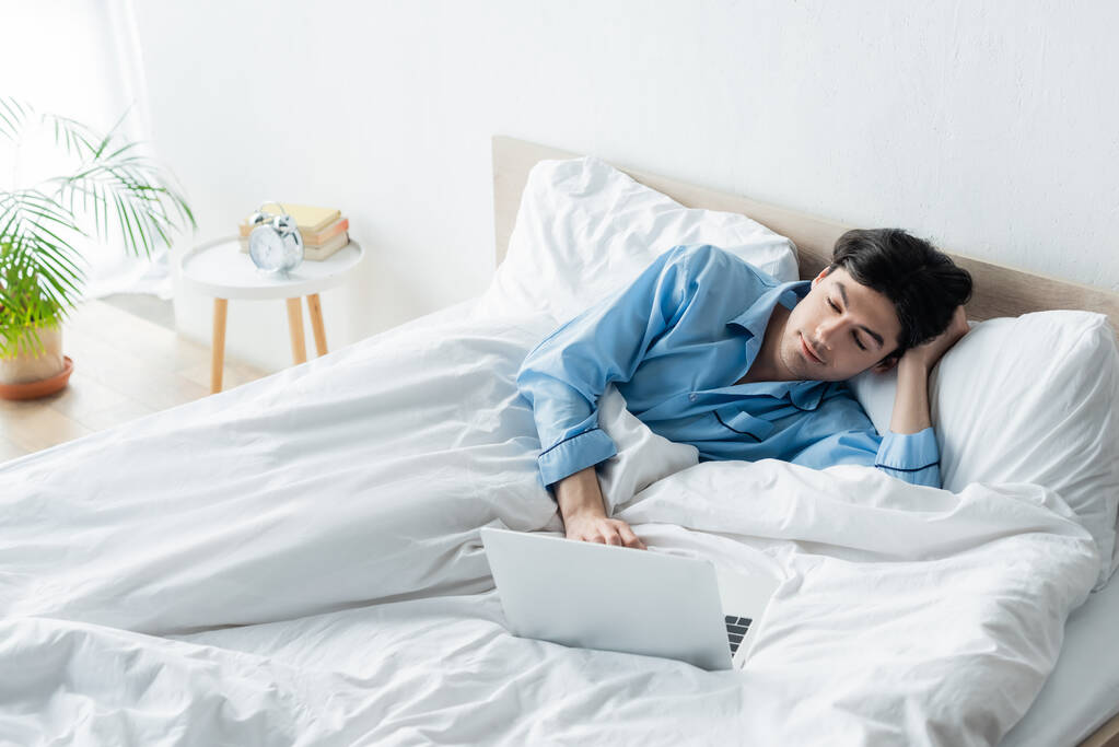 hombre positivo en pijama azul viendo la película en el ordenador portátil en la cama - Foto, Imagen