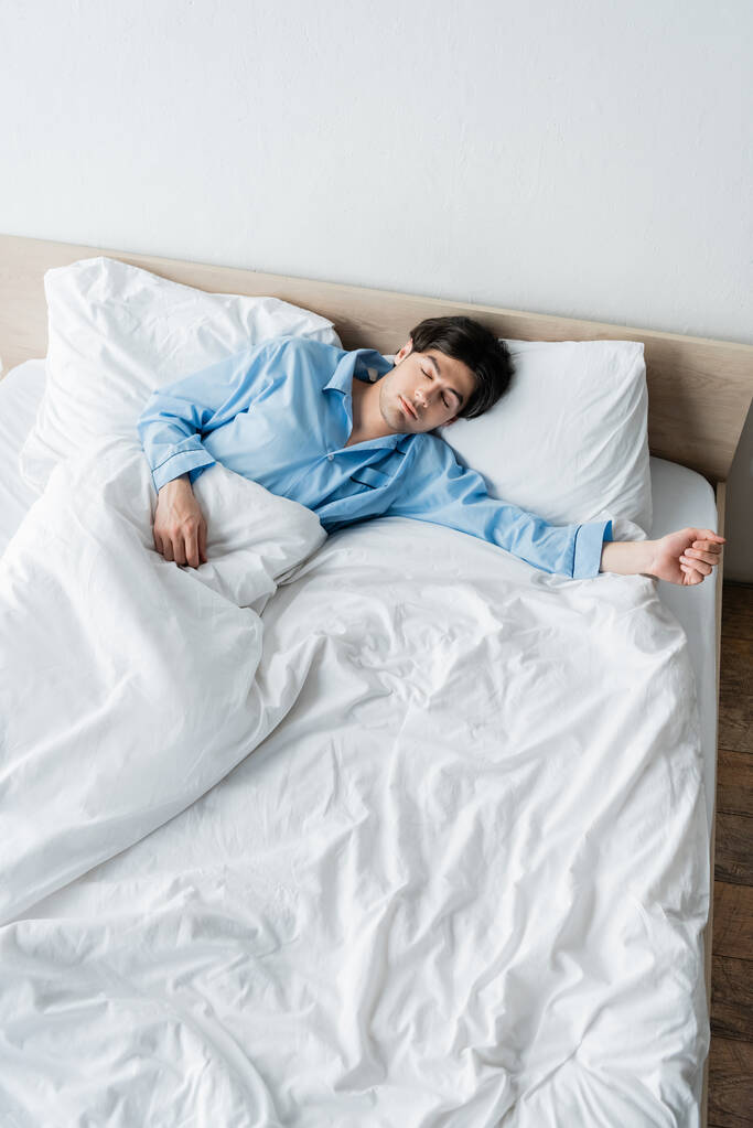 vysoký úhel pohledu na mladého muže v modrém pyžamu spí na bílém lůžku - Fotografie, Obrázek