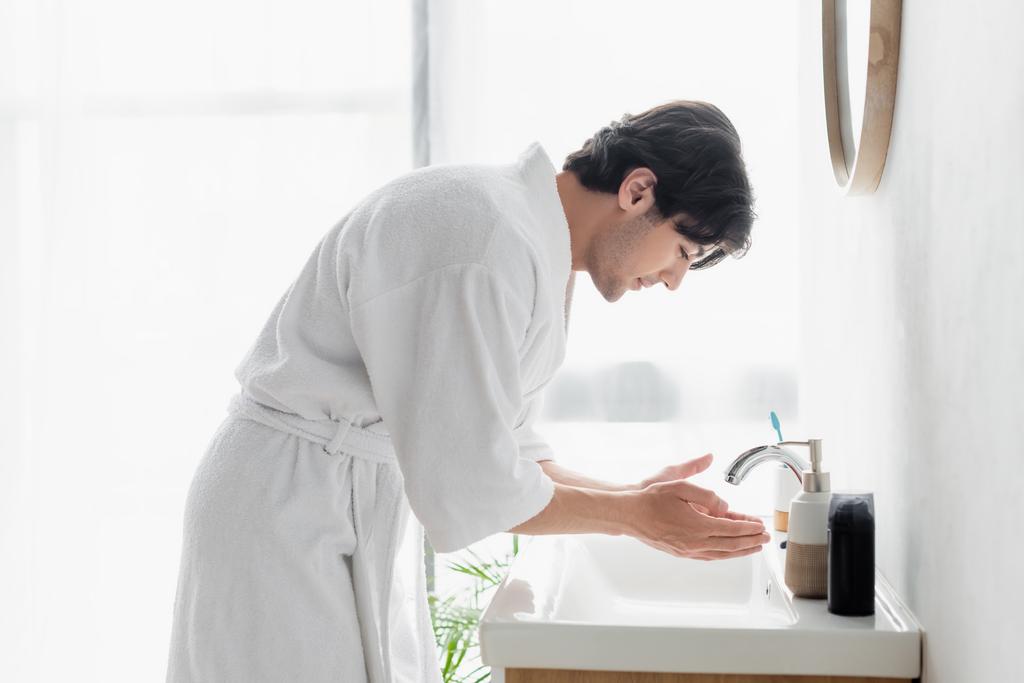 side view of young man in white bathrobe near sink and toiletries in bathroom - Photo, Image