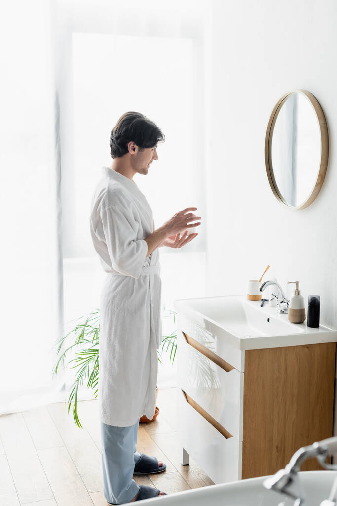 vue pleine longueur de l'homme en peignoir blanc tenant crème cosmétique près du miroir et articles de toilette sur l'évier - Photo, image