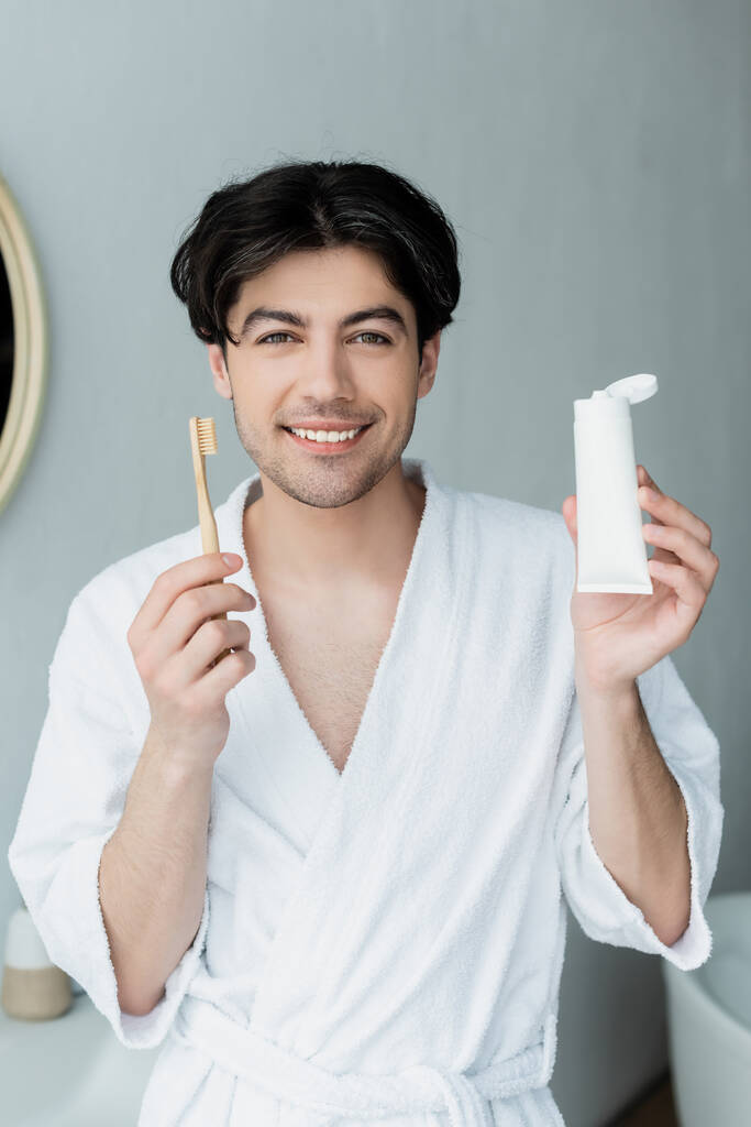 gelukkig man met tandenborstel en buis van tandpasta kijken in de camera in de badkamer - Foto, afbeelding