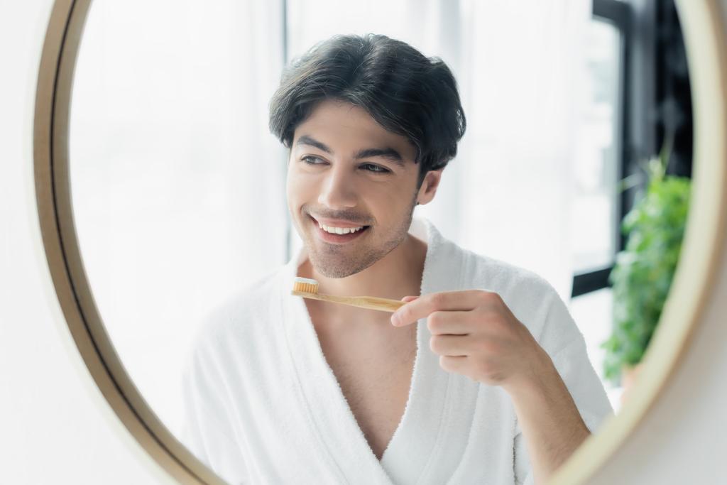 uomo allegro in accappatoio bianco in piedi vicino allo specchio del bagno con spazzolino da denti - Foto, immagini