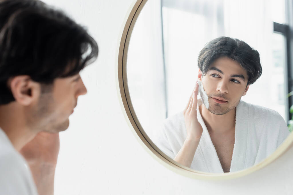 брюнетка мужчина смотрит в зеркало ванной комнаты при нанесении пены для бритья - Фото, изображение