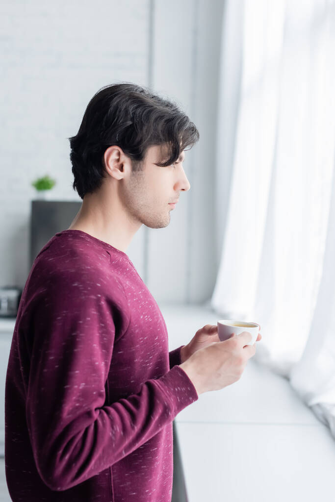 Вид сбоку молодого брюнета, стоящего с чашкой кофе у окна  - Фото, изображение