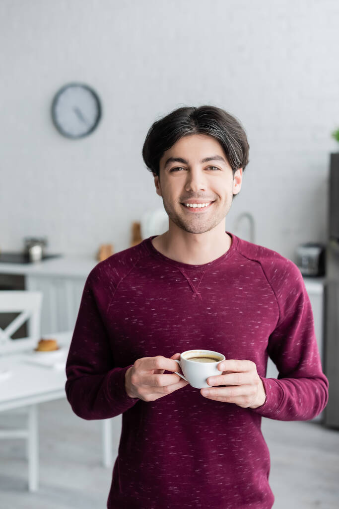 szczęśliwy młody człowiek trzyma filiżankę kawy patrząc na aparat fotograficzny w kuchni - Zdjęcie, obraz