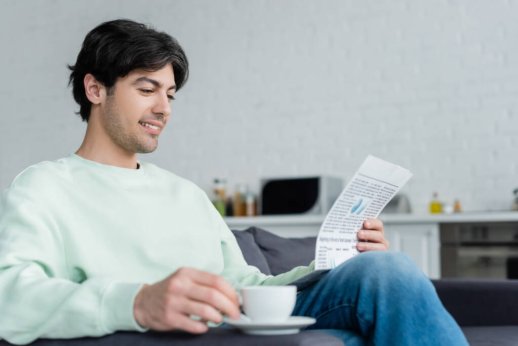 улыбающийся человек читает газету возле размытой чашки кофе на диване - Фото, изображение