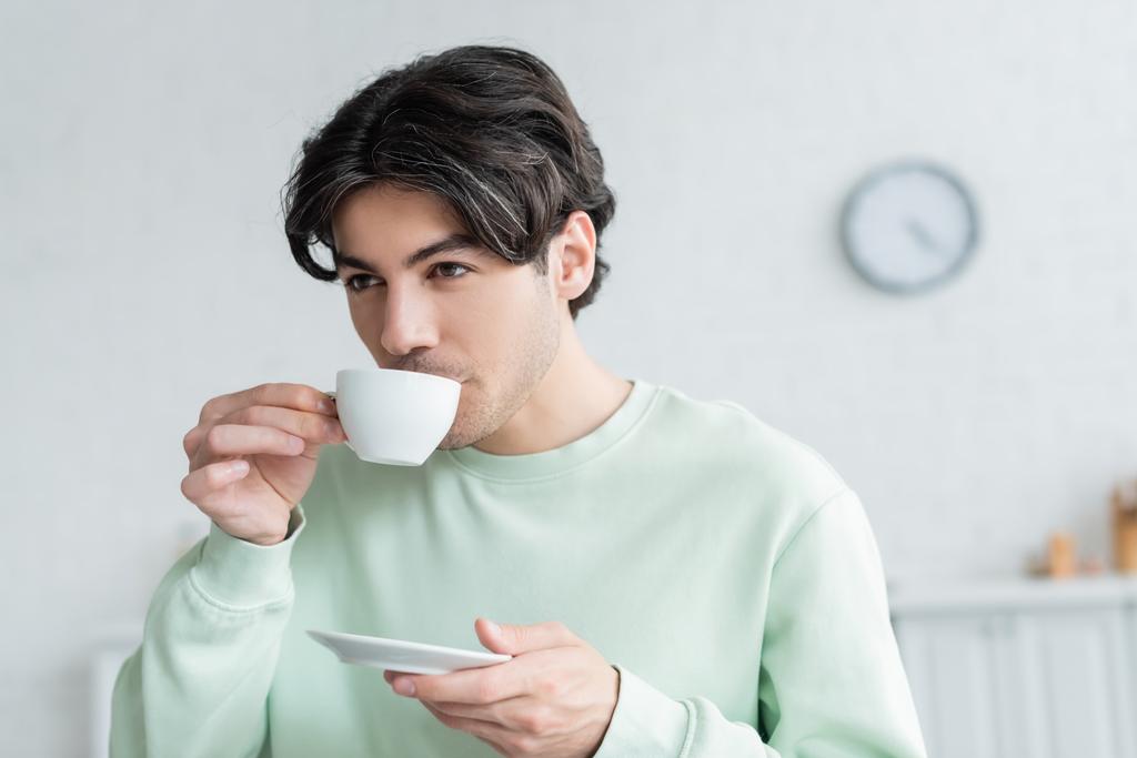 νεαρός μελαχρινός άνδρας κρατώντας πιατάκι, ενώ πίνοντας πρωινό καφέ - Φωτογραφία, εικόνα