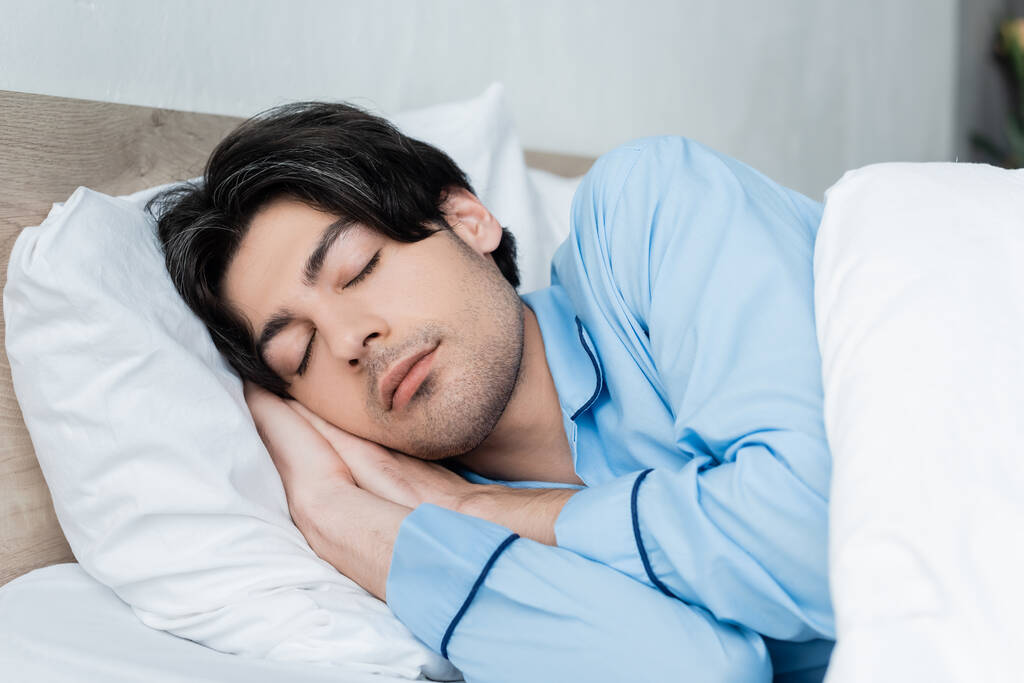 Mavi pijamalı genç esmer adam beyaz yatakta uyuyor. - Fotoğraf, Görsel