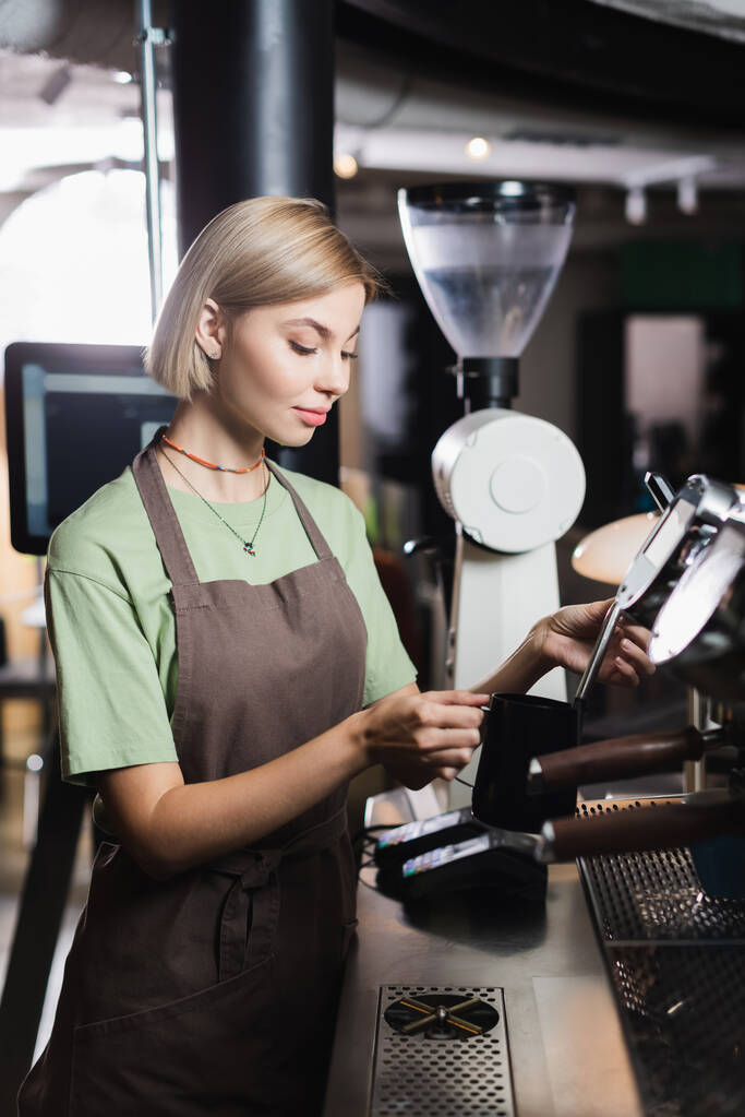 Fiatal barista kötényben kezében gőzgép és tejes kancsó kávézóban  - Fotó, kép
