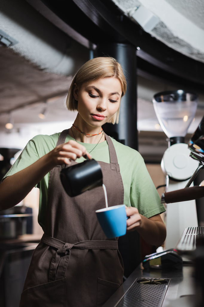 Блондинка-бариста в фартуке наливает молоко в чашку возле кофеварки в кафе  - Фото, изображение