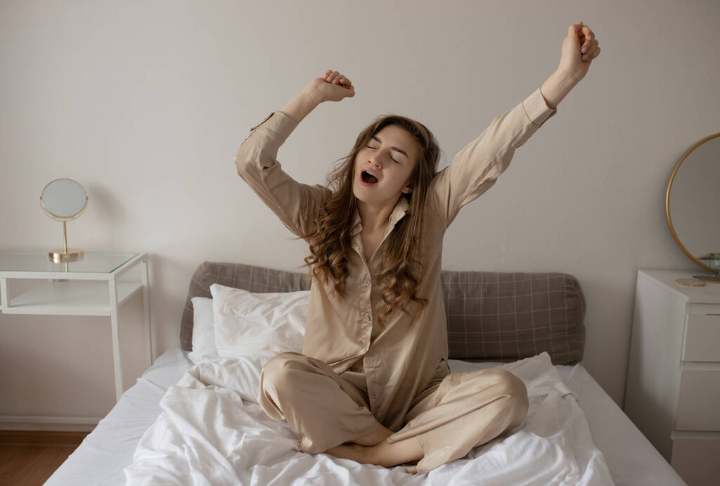 Une jeune fille en pyjama beige est penchée au lit. La fille bâille le matin assise au lit. Mise au point sélective - Photo, image