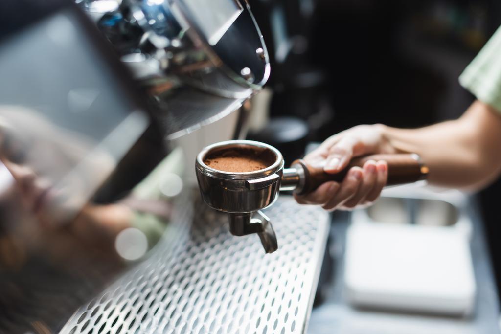 Обрезанный вид на бариста, держащего портативный фильтр с кофе возле размытой кофеварки в кафе - Фото, изображение
