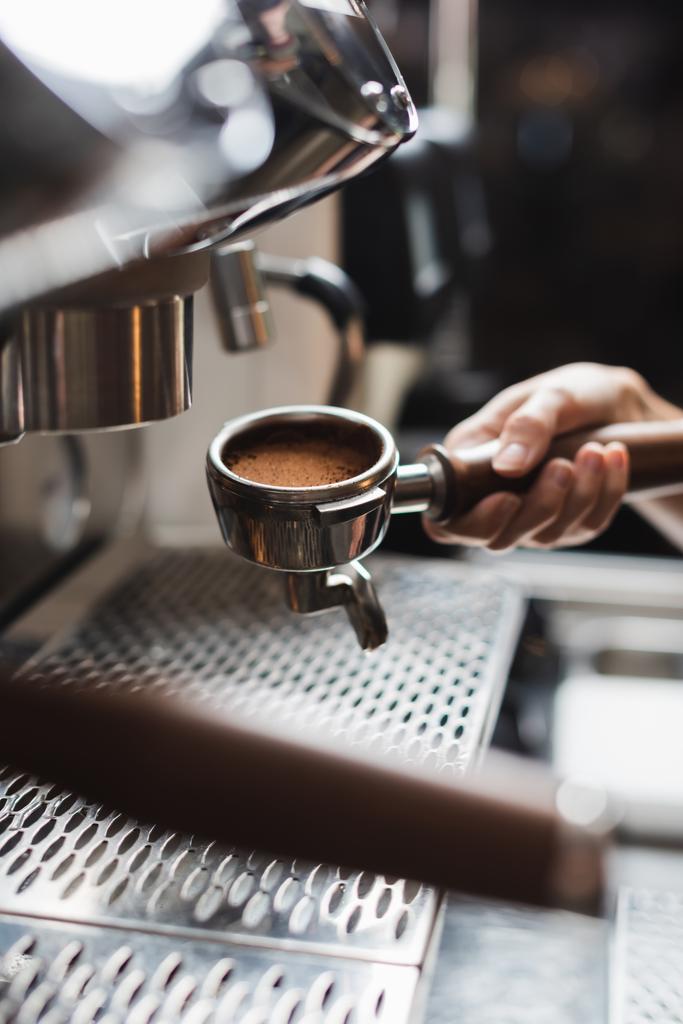 Ausgeschnittene Ansicht des Barista mit Portafilter und Kaffee in der Nähe einer verschwommenen Kaffeemaschine im Vordergrund im Café  - Foto, Bild
