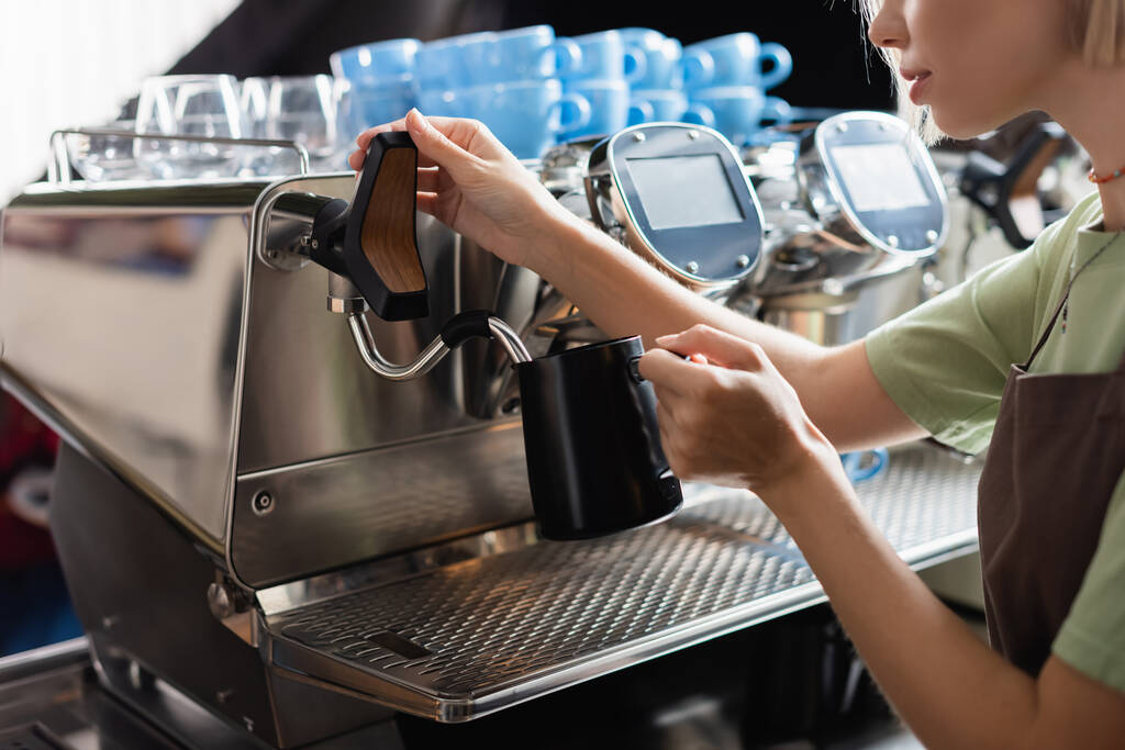 Vágott kilátás barista kezében tejes kancsó mellett kávéfőző gőzölgő pálcával kávézóban  - Fotó, kép