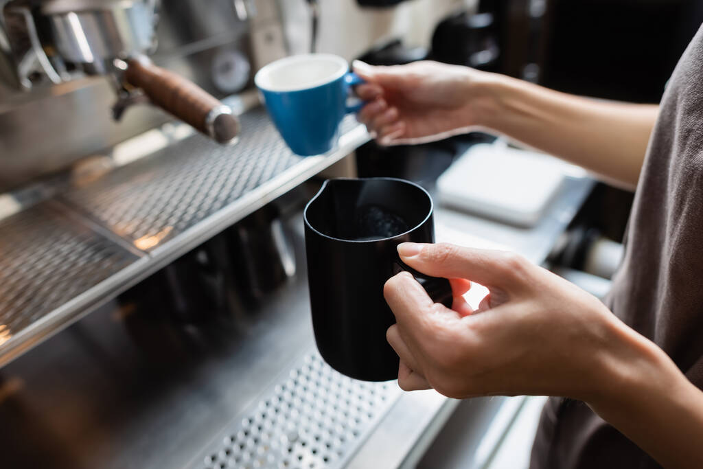 Vista cortada de barista segurando jarro de leite e xícara borrada perto da máquina de café no café  - Foto, Imagem