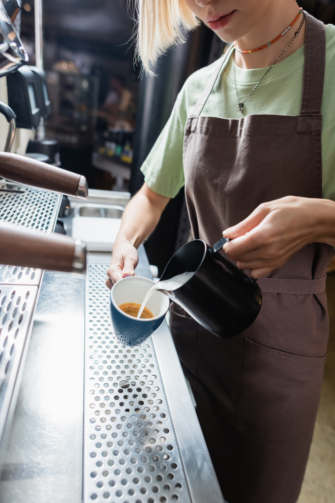 Vágott kilátás barista kötényben öntés tej csésze kávé mellett kávéfőző gép kávézóban  - Fotó, kép