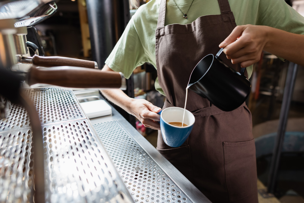 Vista recortada de barista en delantal vertiendo leche de jarra en taza en la cafetería  - Foto, Imagen