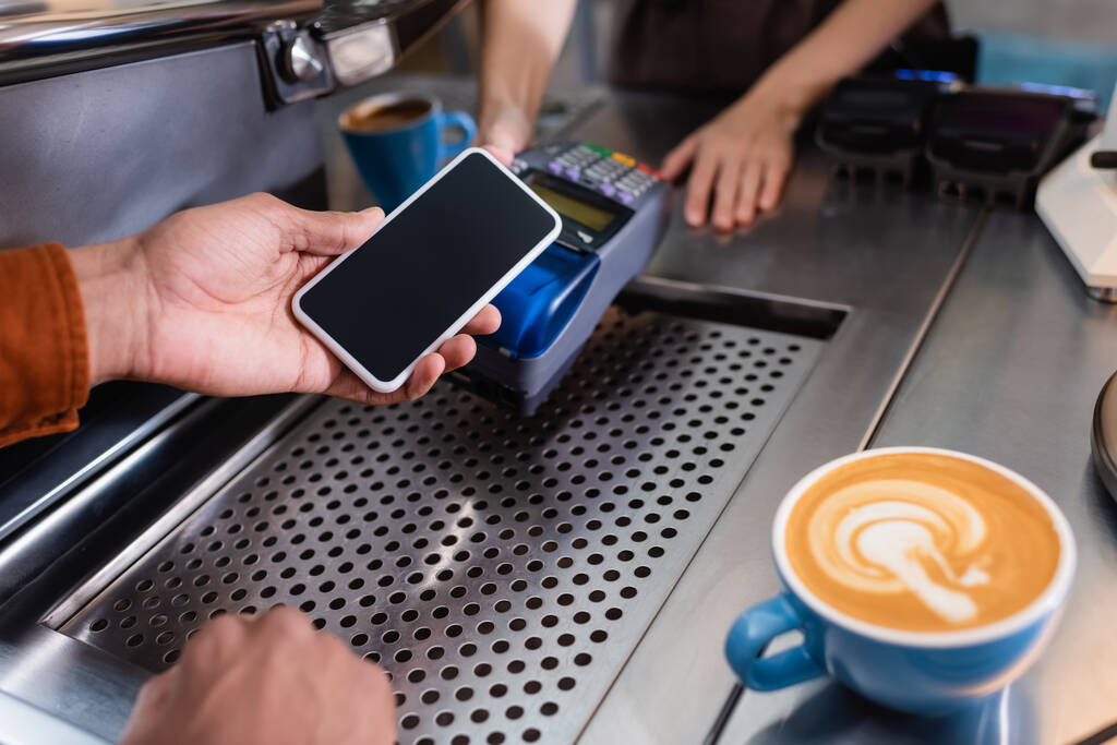Vista recortada del hombre afroamericano sosteniendo teléfono celular cerca de barista con terminal de pago y café en la cafetería  - Foto, imagen